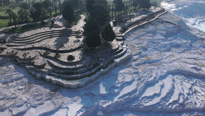 Aerial drone footage of the white travertine terraces of Pamukkale in Turkey, filled with natural thermal water pools. The bright mineral formations create a striking contrast with the surrounding landscape under soft sunlight. This unique geological site, formed by calcium-rich hot springs, is one of Turkey’s most iconic natural landmarks and a UNESCO World Heritage location, ideal for travel, tourism, documentary, and nature projects.