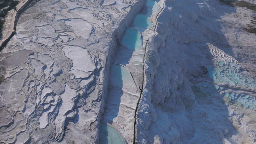 Aerial drone footage of the white travertine terraces of Pamukkale in Turkey, filled with natural thermal water pools. The bright mineral formations create a striking contrast with the surrounding landscape under soft sunlight. This unique geological site, formed by calcium-rich hot springs, is one of Turkey’s most iconic natural landmarks and a UNESCO World Heritage location, ideal for travel, tourism, documentary, and nature projects.