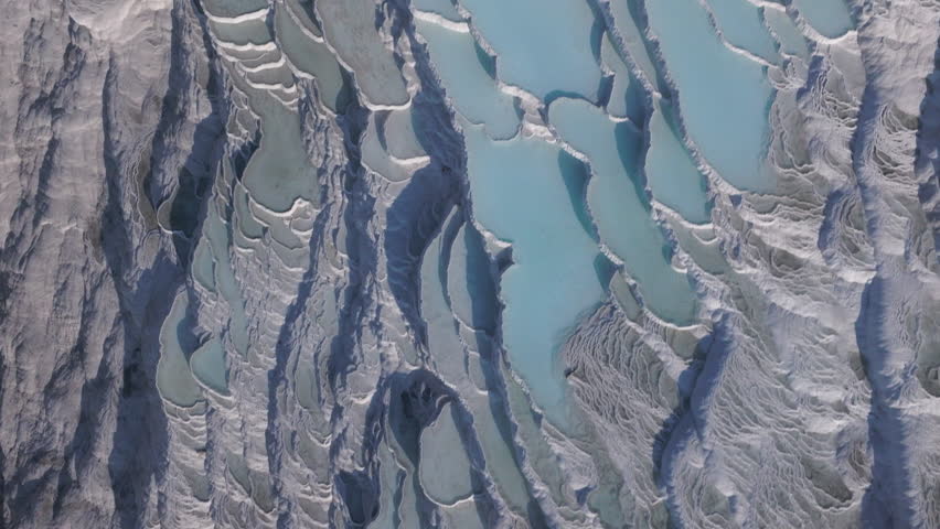 Aerial drone footage of the white travertine terraces of Pamukkale in Turkey, filled with natural thermal water pools. The bright mineral formations create a striking contrast with the surrounding landscape under soft sunlight. This unique geological site, formed by calcium-rich hot springs, is one of Turkey’s most iconic natural landmarks and a UNESCO World Heritage location, ideal for travel, tourism, documentary, and nature projects.