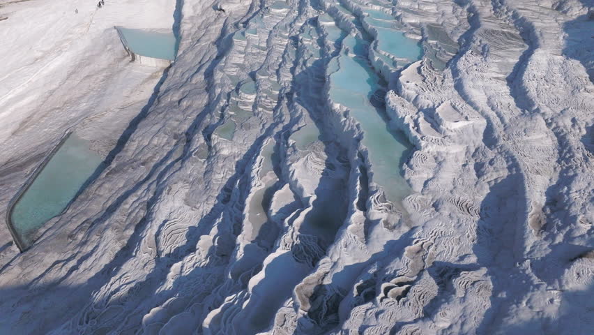 Aerial drone footage of the white travertine terraces of Pamukkale in Turkey, filled with natural thermal water pools. The bright mineral formations create a striking contrast with the surrounding landscape under soft sunlight. This unique geological site, formed by calcium-rich hot springs, is one of Turkey’s most iconic natural landmarks and a UNESCO World Heritage location, ideal for travel, tourism, documentary, and nature projects.