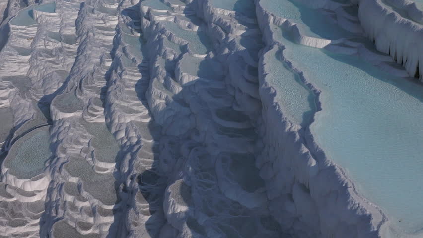 Aerial drone footage of the white travertine terraces of Pamukkale in Turkey, filled with natural thermal water pools. The bright mineral formations create a striking contrast with the surrounding landscape under soft sunlight. This unique geological site, formed by calcium-rich hot springs, is one of Turkey’s most iconic natural landmarks and a UNESCO World Heritage location, ideal for travel, tourism, documentary, and nature projects.