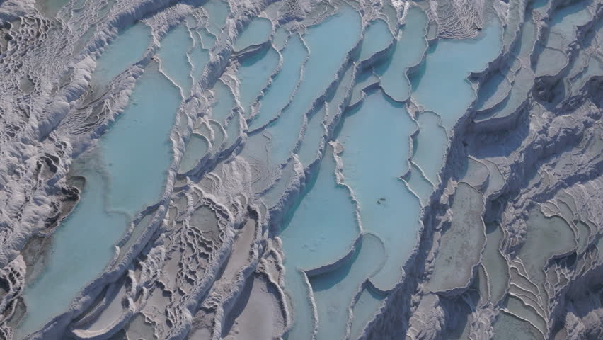 Aerial drone footage of the white travertine terraces of Pamukkale in Turkey, filled with natural thermal water pools. The bright mineral formations create a striking contrast with the surrounding landscape under soft sunlight. This unique geological site, formed by calcium-rich hot springs, is one of Turkey’s most iconic natural landmarks and a UNESCO World Heritage location, ideal for travel, tourism, documentary, and nature projects.