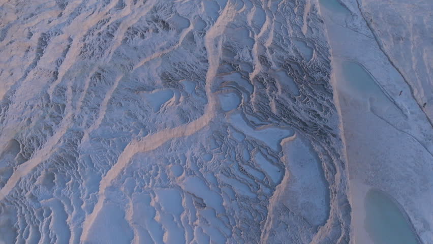 Aerial drone footage of the white travertine terraces of Pamukkale in Turkey, filled with natural thermal water pools. The bright mineral formations create a striking contrast with the surrounding landscape under soft sunlight. This unique geological site, formed by calcium-rich hot springs, is one of Turkey’s most iconic natural landmarks and a UNESCO World Heritage location, ideal for travel, tourism, documentary, and nature projects.