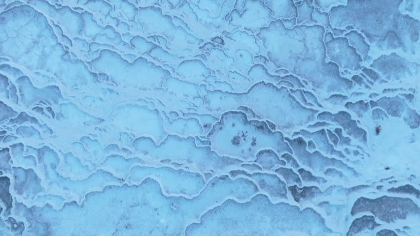 Aerial drone footage of the white travertine terraces of Pamukkale in Turkey, filled with natural thermal water pools. The bright mineral formations create a striking contrast with the surrounding landscape under soft sunlight. This unique geological site, formed by calcium-rich hot springs, is one of Turkey’s most iconic natural landmarks and a UNESCO World Heritage location, ideal for travel, tourism, documentary, and nature projects.