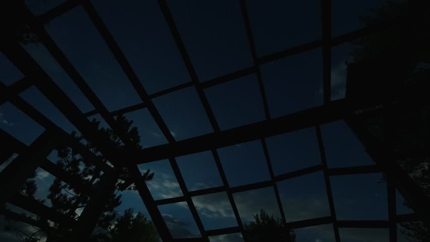Moon passes over an abandoned greenhouse building.  Only the framework remains.  Partly cloudy night sky with stars and airplanes passing by.  Timelapse nightime