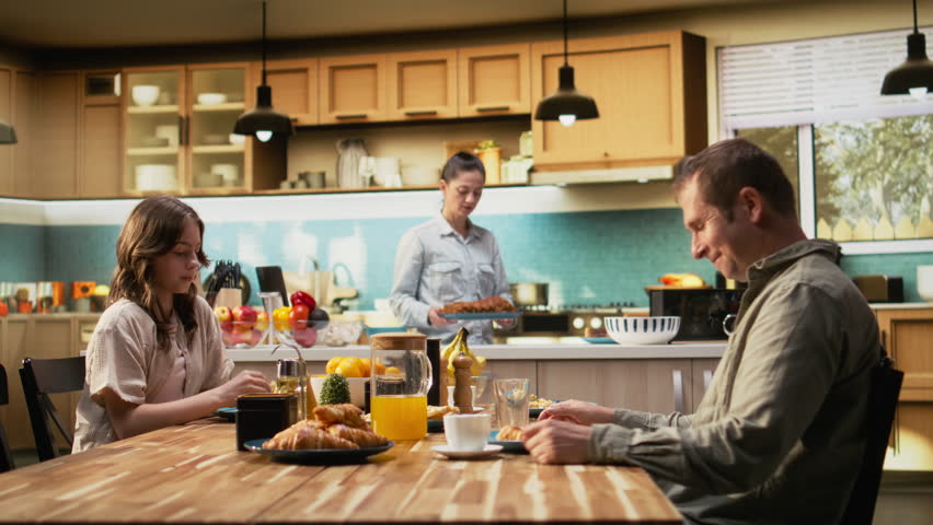 Family with a pre-teen girl serving breakfast together in a bright kitchen, parents pour orange juice and daughter enjoys eggs and toast. Cozy scene captures love and bonding time. Camera A.