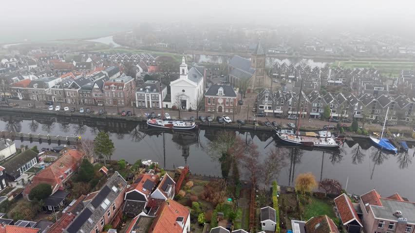 Aerial view of foggy Dutch canal town gabled brick homes, docked boats, and tall church tower form a tranquil, symmetrical waterfront scene softened by mist.