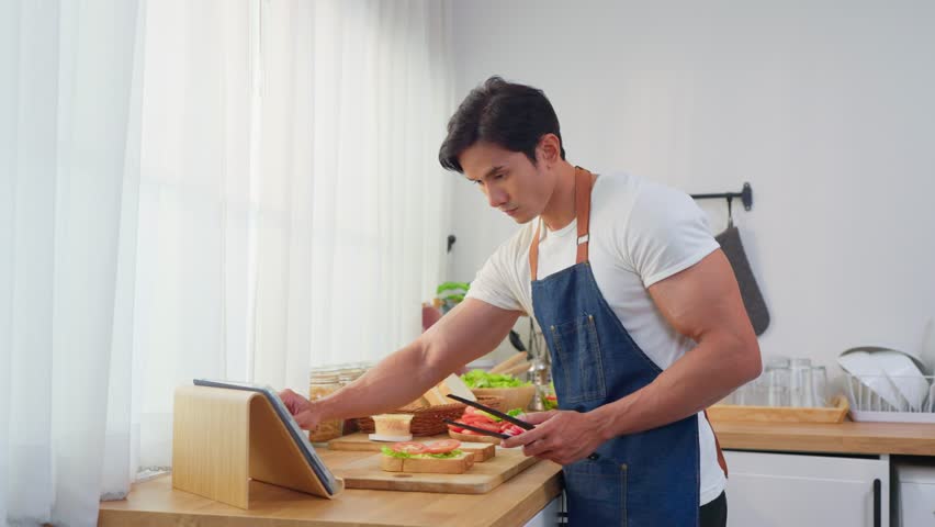 Asian man cooking healthy foods in cozy kitchen in morning at home. Attractive young male feeling happy and relax, enjoy preparing ingredients and making healthy sandwich bread for breakfast in house.
