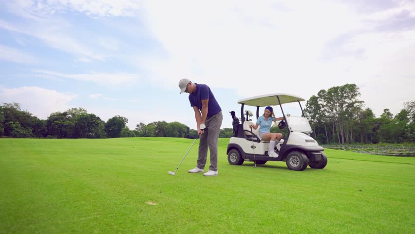 Asian man and woman golfers swing clubs together on scenic golf course. Attractive people enjoy fun game outdoor recreation and light exercise while practicing under the guidance of trainer in course.
