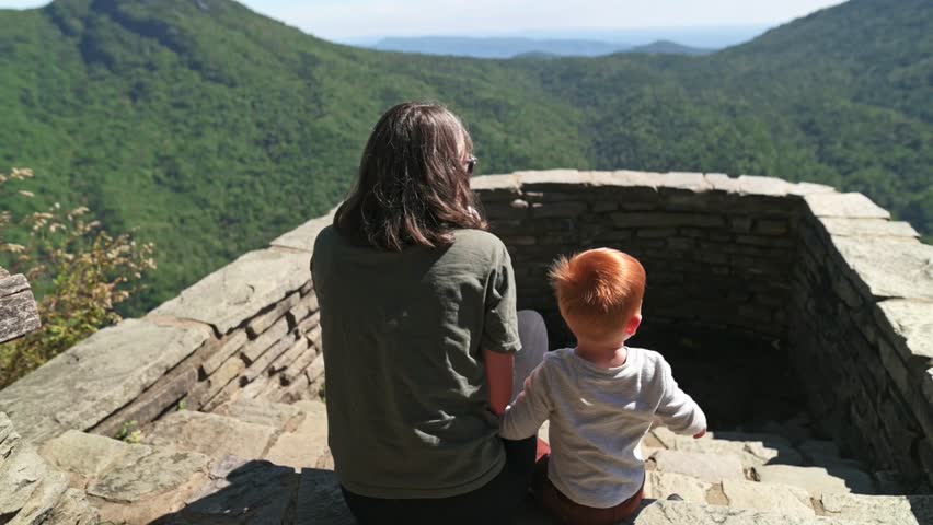 White Parent Redhead Child Seated On Stone Lookout Facing Sweeping Mountain Valley, Gentle Breeze Through Trees, Peaceful Summer Afternoon, Child Pointing Toward Distant Ridge, Tactile Stone