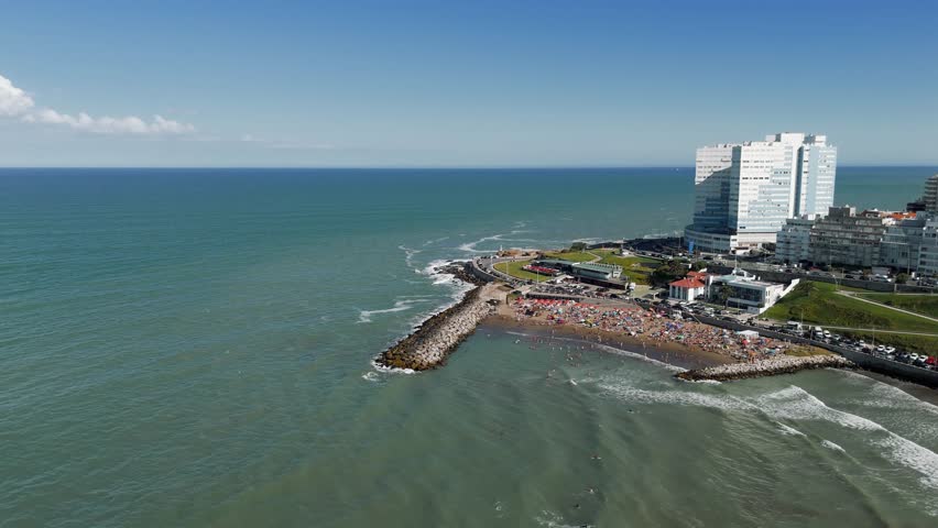 Aerial view of Cabo Corrientes beach in Mar del Plata, Argentina, with people enjoying a sunny summer day. The camera performs an approach, push in, tilt down, and rotate movement