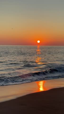 A cinematic sunset over the ocean, showing the glowing orange sun slowly descending toward the horizon. Gentle sea waves reflect warm golden and red tones, while the sky transitions smoothly from deep orange to soft dusk colors, creating a calm, peaceful, and atmospheric seascape ideal for nature, travel, and relaxation themes.