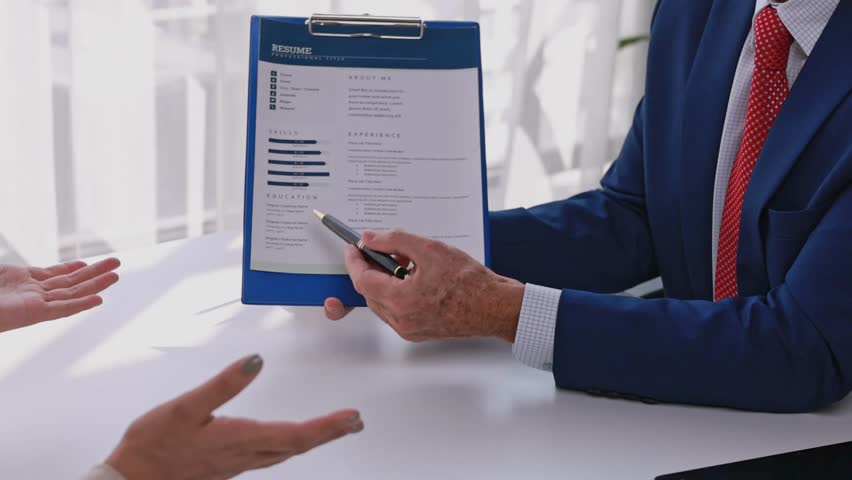 Recruiter pointing at a resume on a clipboard while talking to a job applicant in an office