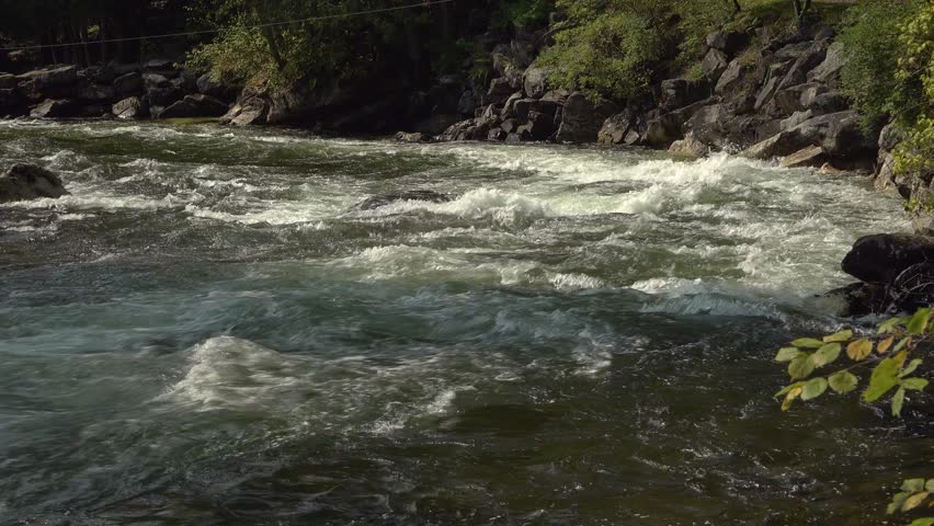Small rapids on the bend of a sunny river in a forest with a rocky shoreline during the summer.