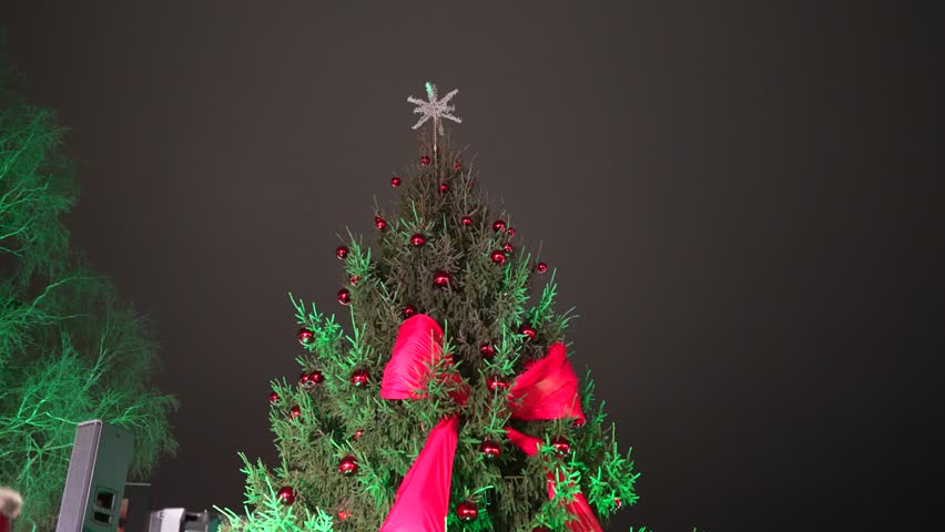 A beautifully decorated Christmas tree stands tall with vibrant lights, red bows, and sparkling ornaments, creating a joyful holiday scene under the night sky