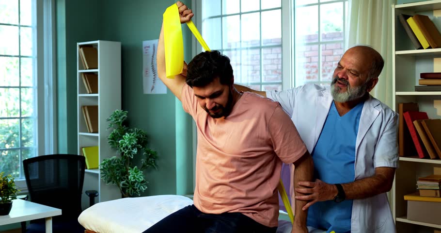 Indian physiotherapist teaching patient resistance band exercises for shoulder and tricep as senior male therapist assists strength training on clinic bed inside modern Indian clinic