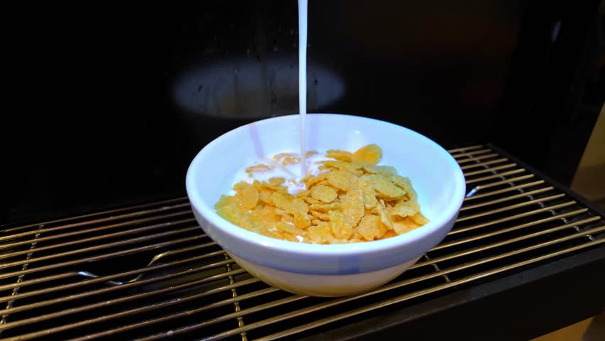 Milk being poured into cornflakes cereal in white bowl during breakfast preparation in home kitchen