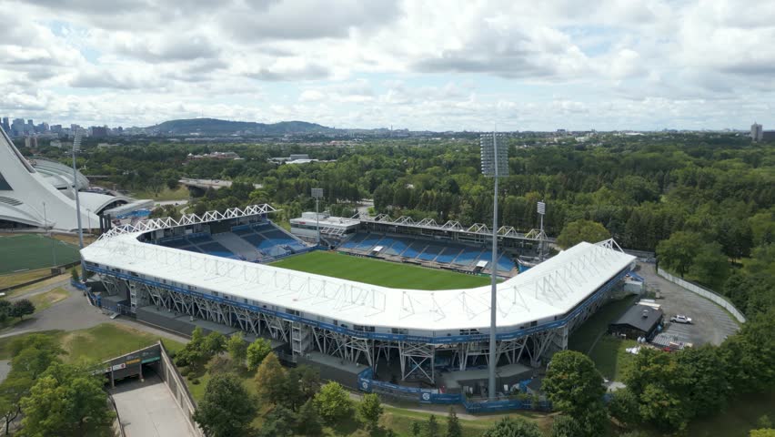 Aerial of Montreal olympic stadium with curved roof, green parkland, urban skyline backdrop, establishing orbit