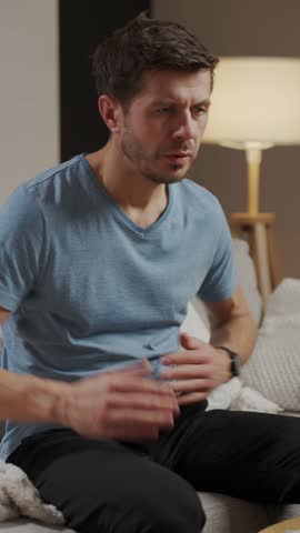 Man sitting on sofa in living room holding stomach and suffering from abdominal pain or cramps. Person suffering from health issues, digestion problems or food poisoning at home
