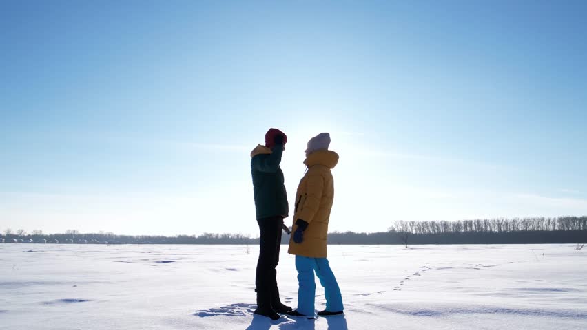 Young couple standing in a snowy winter field adjusting each others hat and hood. Warm care, support and affection in cold weather. Minimal winter landscape with copy space, backlit scene.