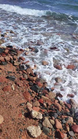 Waves continuously crash against the rocky shoreline creating foam and splashes over pebbles and sand along the beach