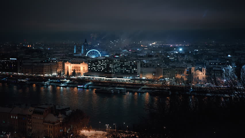 New Year fireworks light up Budapest along the Danube riverbank, revealing illuminated bridges, historic architecture, and vibrant city lights. Festive aerial night view of Hungary’s capital.