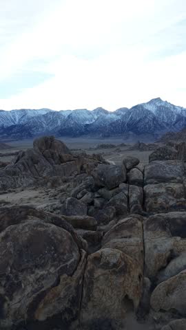 Vertical 4K drone footage slowly flying through Alabama Hills, California, showcasing rugged boulders, snowy-capped Eastern Sierra mountains, and expansive natural desert landscapes.