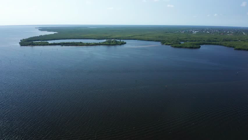Calm ocean water surrounds green mangrove islands along the Florida coast, with a distant residential area visible on the horizon under a bright, partly cloudy sky.