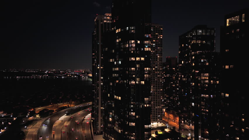 Night aerial view of Chicago, Illinois skyline with illuminated residential and business towers and glowing highway light trails, deep contrast, urban density, vibrant metropolitan atmosphere.