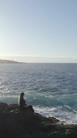 Young woman with long blonde hair sitting on a rocky shore looking at the sea