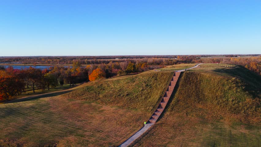 Drone footage zooming out from Monk’s Mound at Cahokia Mounds State Historic Site. The shot reveals the surrounding landscape as colorful autumn trees contrast with a bright blue sky on a sunny day.