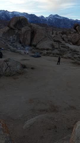 Aerial 4K vertical drone shot rising above a campsite with a burning campfire beside a tent and a lone camper in the rugged Alabama Hills rock landscape at golden hour