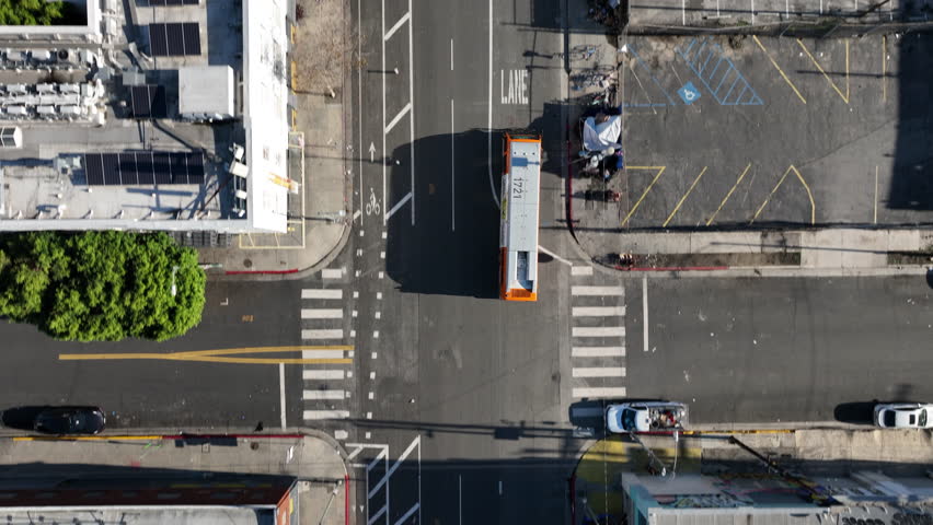 Top-down aerial drone view of a Los Angeles street showing a bus in traffic and nearby homeless encampments, documenting urban transportation, street life, and social conditions.