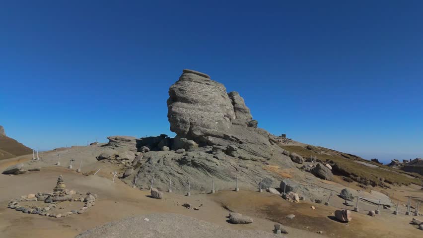 Slow 4K aerial drone push-in shot focusing on the intricate details of the Sphinx (Sfinxul) rock formation in the Bucegi Mountains, Romania. The camera moves intimately close to the wind-eroded conglomerate surface, revealing the rough textures and the iconic profile against a clear deep blue sky.