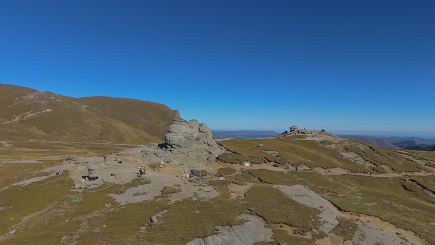 Wide 4K aerial drone orbit capturing the entire Babele and Sphinx tourist complex in the Bucegi Mountains, Romania. The distant perspective showcases the spatial layout of these iconic wind-eroded conglomerate formations, the nearby cable car station, and the winding footpaths traversing the alpine plateau. The scene emphasizes the vast scale of the high-altitude landscape covered in Nardus stricta grass under a sunny blue sky, offering a comprehensive view of this popular Carpathian landmark.