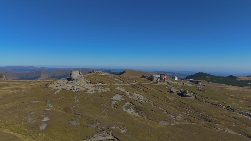 Wide 4K aerial drone orbit capturing the entire Babele and Sphinx tourist complex in the Bucegi Mountains, Romania. The distant perspective showcases the spatial layout of these iconic wind-eroded conglomerate formations, the nearby cable car station, and the winding footpaths traversing the alpine plateau. The scene emphasizes the vast scale of the high-altitude landscape covered in Nardus stricta grass under a sunny blue sky, offering a comprehensive view of this popular Carpathian landmark.