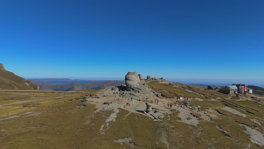 Immersive 4K aerial drone footage moving in a straight line towards the iconic Sphinx (Sfinxul) rock formation in the Bucegi Mountains, Romania. As the camera approaches the weathered conglomerate face, it ascends slightly to execute a smooth flyover, revealing the top texture of the megalith and the vast alpine plateau covered in dry Nardus stricta grass stretching out behind it. The shot captures the sheer scale of the geological landmark against a backdrop of intense blue sky.