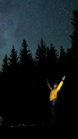 Human silhouette beneath star trails above dark conifer woodland, Explorer lifts arms skyward experiencing cosmic motion during silent nocturnal landscape, vertical footage
