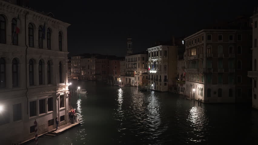 Grand Canal in Venice at night with moving water taxis and boats. Historic buildings and lights reflect on dark water. People stand on a small pier during busy evening in Italian city.