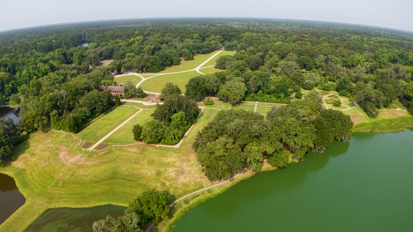 Aerial footage of a landscaped park in Charleston, South Carolina shows manicured lawns, a large pond and a narrow walkway crossing the water, surrounded by lush trees and forest.