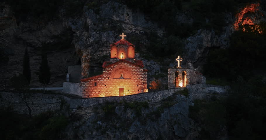 Cinematic drone pull-back shot revealing an illuminated mountain church carved into rocky cliffs above Berat, Albania at blue hour