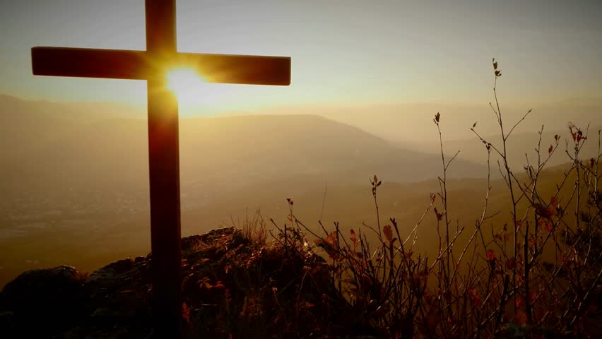 Silhouette of a Christian cross positioned on a mountain or hilltop, set against a glowing sunrise 