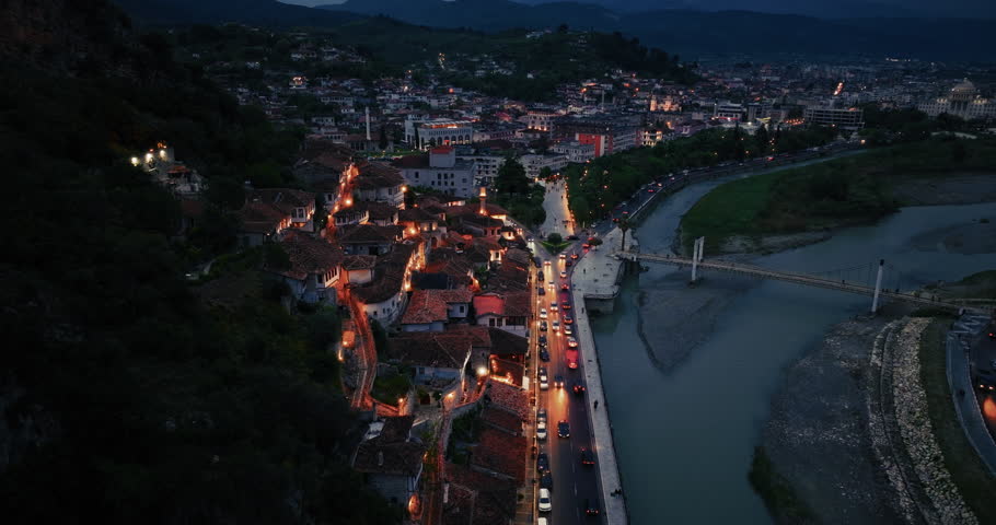 Cinematic aerial view of Berat old town at blue hour with glowing historic houses, river promenade, and evening traffic flowing through the city