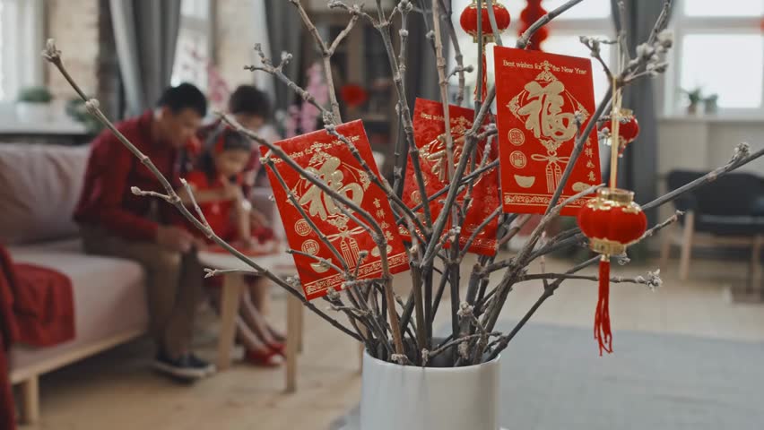 Close-up of postcards and Chinese New Year decorations on branches, while an unrecognizable Asian family with child creates festive crafts in background.
