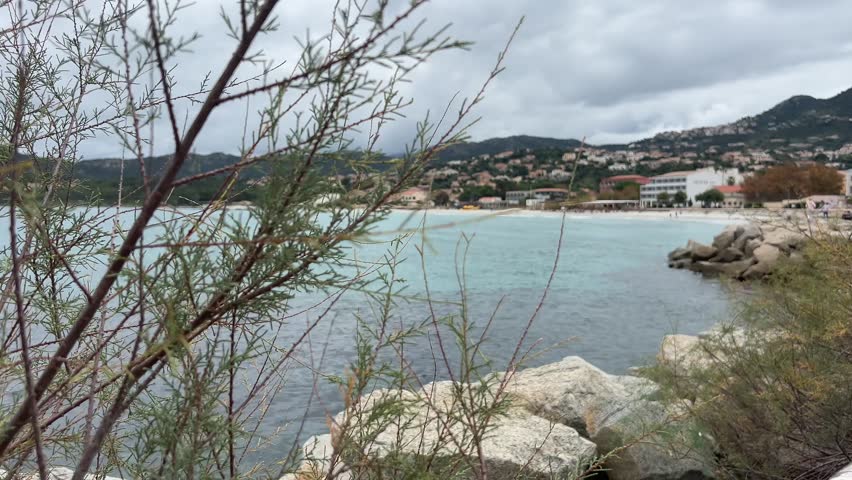Scenic coastal view of L’Île-Rousse in Corsica, featuring turquoise sea, rocky shoreline, and hillside town under a cloudy sky.