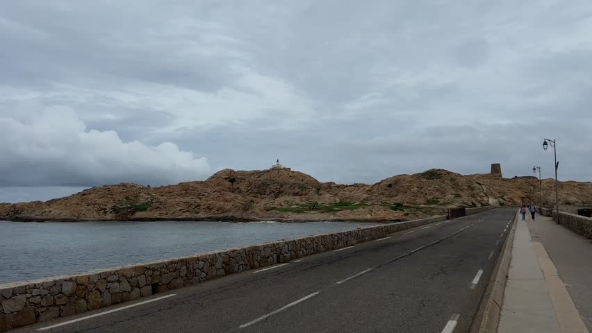 Quiet coastal road along the shoreline in Corsica, France, with rocky headland, calm sea, and cloudy sky creating a peaceful Mediterranean landscape.