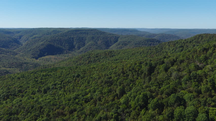 Aerial ascend establishing rolling Ozark highlands covered in dense mixed hardwood forest under clear autumn sky, Arkansas