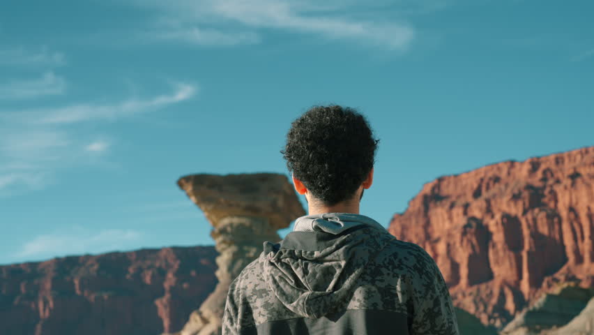 Rear view of a male tourist in a camo hoodie standing still and admiring El Hongo rock formation in Ischigualasto, San Juan, contrasting unique geological shapes against deep blue sky.