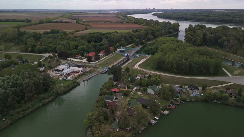 Orbiting aerial view of a sluice along the Ráckevei Danube River in Tass, Hungary, surrounded by a tranquil residential area, lush greenery, and scenic rural landscapes.