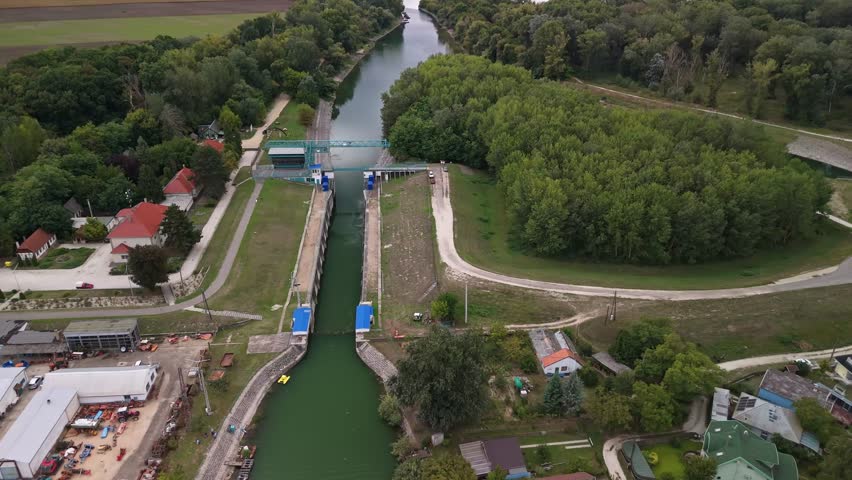 Tilt-down aerial view of a sluice gate on the Ráckevei Danube in Tass, Hungary, surrounded by lush greenery, rural buildings, and scenic countryside, showcasing water management and natural beauty.
